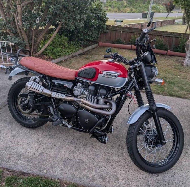May include: A red and silver Triumph motorcycle with a brown leather seat and a black exhaust pipe. The motorcycle has a black front fender and a black rear fender. The motorcycle is parked on a gray driveway.