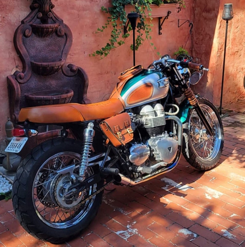 K&ouml;nnte beinhalten: Ein gr&uuml;n-silbernes Motorrad mit braunen Ledersitzen und Satteltaschen. Das Motorrad hat einen verchromten Auspuff und R&auml;der. Das Motorrad steht vor einer Backsteinmauer mit einem Springbrunnen.