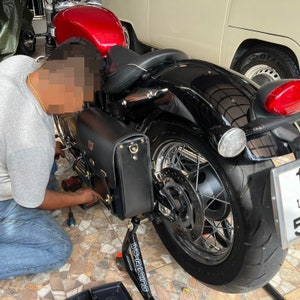 May include: A black motorcycle with a red gas tank and a black leather saddlebag. The motorcycle is on a stand and has a license plate that says "155".