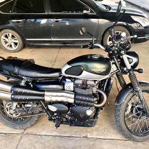 May include: A Triumph Scrambler motorcycle with a green and silver fuel tank, black seat, and dual exhaust pipes. The motorcycle has black tires and a black frame. The word "Triumph" is visible on the fuel tank.