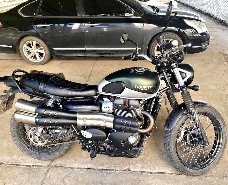 May include: A Triumph Scrambler motorcycle with a green and silver fuel tank, black seat, and dual exhaust pipes. The motorcycle has black tires and a black frame. The word "Triumph" is visible on the fuel tank.