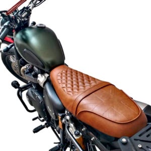 May include: A motorcycle seat with a brown, diamond-stitched leather seat. The seat is attached to a dark green motorcycle tank and black frame. The handlebars and front wheel are visible in the background.