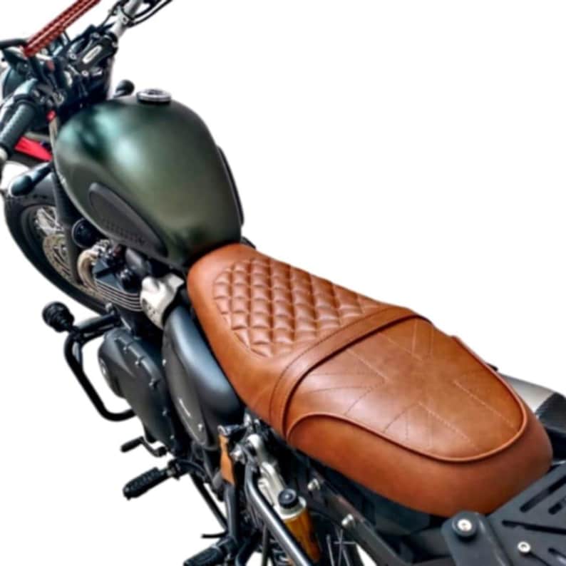 May include: A motorcycle seat with a brown, diamond-stitched leather seat. The seat is attached to a dark green motorcycle tank and black frame. The handlebars and front wheel are visible in the background.