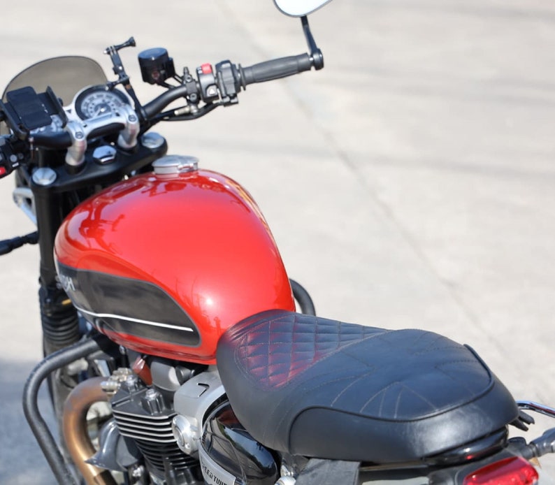 Puede incluir: Una motocicleta roja con un asiento negro y una franja negra y blanca en el tanque de gasolina. La motocicleta tiene un parabrisas negro y un guardabarros trasero cromado.