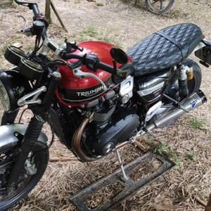 May include: A red Triumph motorcycle with a black quilted seat and a silver exhaust pipe. The motorcycle has a black frame and a silver front fender.