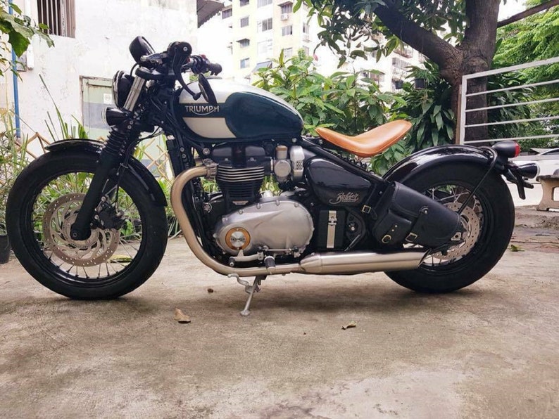 May include: A green and black Triumph motorcycle with a brown leather seat. The motorcycle has a chrome exhaust pipe and a black leather saddlebag.