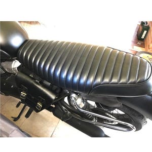 May include: Black motorcycle seat with a ribbed design. The seat is made of a shiny, leather-like material. A black leather saddlebag is attached to the side of the motorcycle. The motorcycle has a black fuel tank and a chrome exhaust pipe.