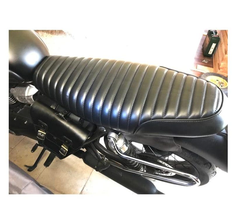 May include: Black motorcycle seat with a ribbed design. The seat is made of a shiny, leather-like material. A black leather saddlebag is attached to the side of the motorcycle. The motorcycle has a black fuel tank and a chrome exhaust pipe.