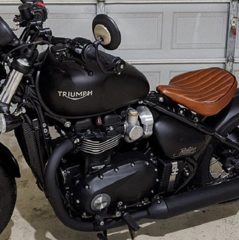 May include: A black Triumph motorcycle with a brown leather seat. The motorcycle has a chrome exhaust pipe and a chrome engine. The motorcycle is parked in a garage.