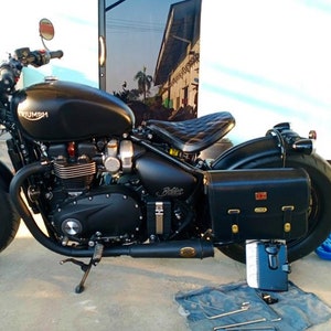 May include: A black Triumph motorcycle with a black leather saddlebag. The motorcycle has a black leather seat and a black exhaust pipe. The motorcycle is parked on a concrete surface.
