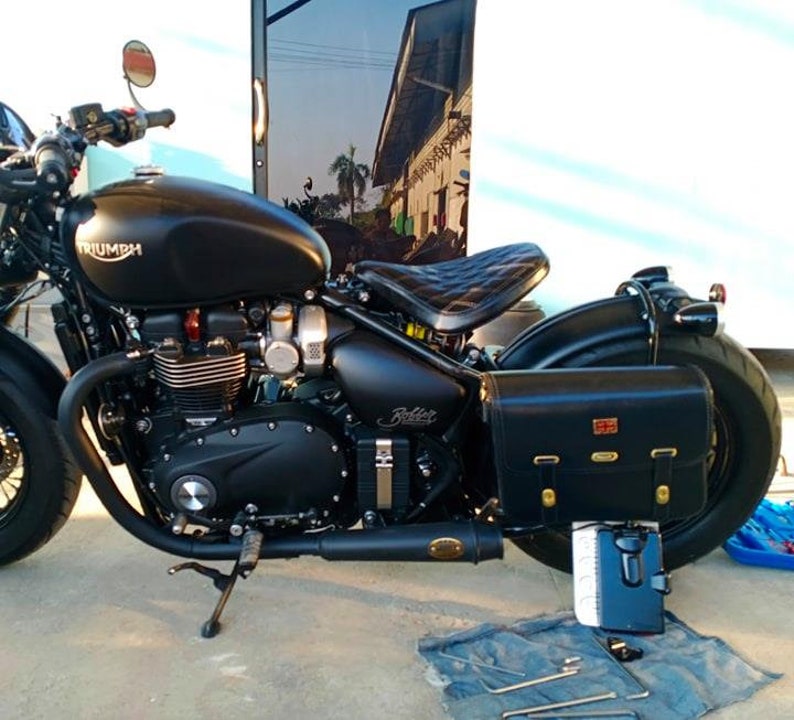 May include: A black Triumph motorcycle with a black leather saddlebag. The motorcycle has a black leather seat and a black exhaust pipe. The motorcycle is parked on a concrete surface.