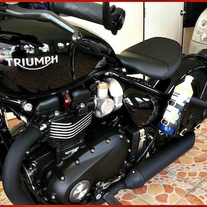 May include: A black Triumph motorcycle with a black leather seat and a black exhaust pipe. The motorcycle has a silver engine and a silver gas tank with the word "TRIUMPH" printed on it. A clear bottle with a blue and yellow label is attached to the side of the motorcycle.
