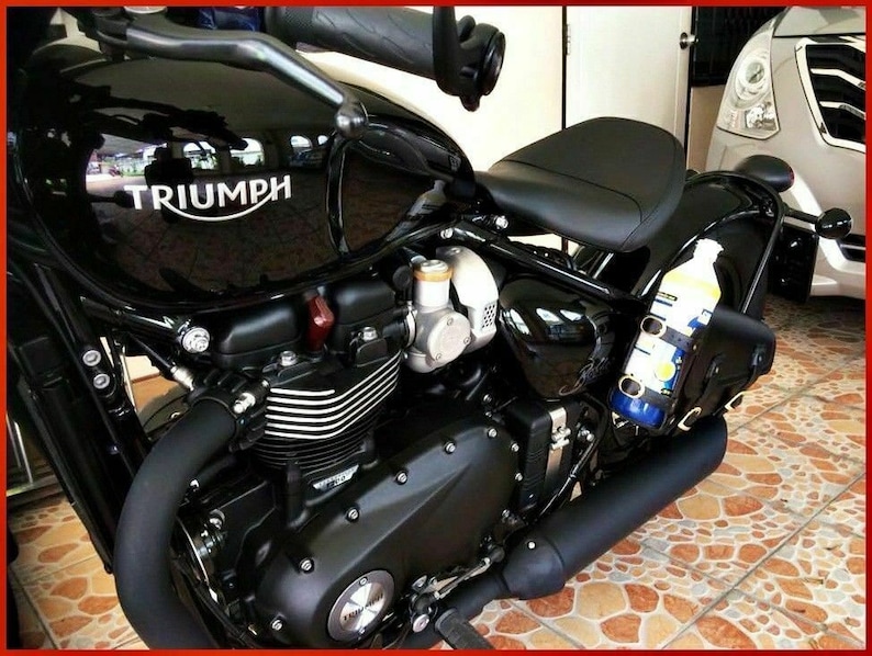 May include: A black Triumph motorcycle with a black leather seat and a black exhaust pipe. The motorcycle has a silver engine and a silver gas tank with the word "TRIUMPH" printed on it. A clear bottle with a blue and yellow label is attached to the side of the motorcycle.