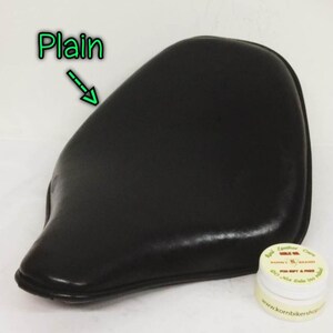 May include: Black leather motorcycle seat with a plain, smooth surface. The seat is curved and has a pointed front. A small jar of leather care product is in the lower right corner of the image. The jar has the text "Real Leather Care Korn's K Brand For Gift & Price"