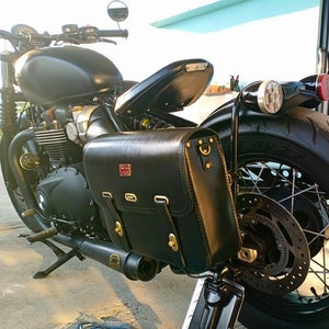 May include: Black motorcycle with a black leather saddlebag with a Union Jack emblem. The motorcycle has a black exhaust pipe and a black rear fender.