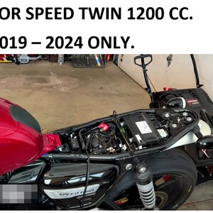 May include: Close-up of a black motorcycle with a red gas tank and a black seat. The motorcycle has a chrome exhaust pipe and a black rear fender. The motorcycle is in a garage and the text "SPEED TWIN" is visible on the side of the motorcycle.