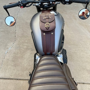 K&ouml;nnte beinhalten: Eine braune Leder-Motorrad-Tanktasche mit einer Schnallenverschlusses und zwei Riemen. Die Tasche ist an einem grauen Motorrad-Benzintank befestigt.