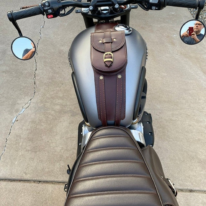 K&ouml;nnte beinhalten: Eine braune Leder-Motorrad-Tanktasche mit einer Schnallenverschlusses und zwei Riemen. Die Tasche ist an einem grauen Motorrad-Benzintank befestigt.