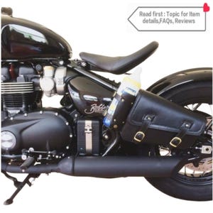 May include: Black motorcycle with a brown leather saddlebag attached to the rear fender. The saddlebag has two buckles and a strap. A clear plastic bottle is secured to the saddlebag with a strap.