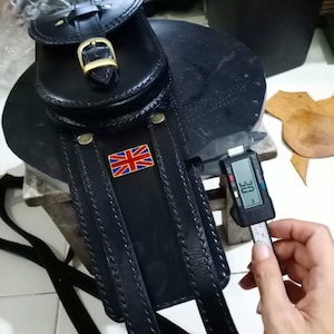 K&ouml;nnte beinhalten: Schwarze Leder-Motorrad-Satteltasche mit einem Union-Jack-Patch und einer Messing-Schnalle. Die Tasche wird mit einem Messschieber gemessen, der 30 mm anzeigt.