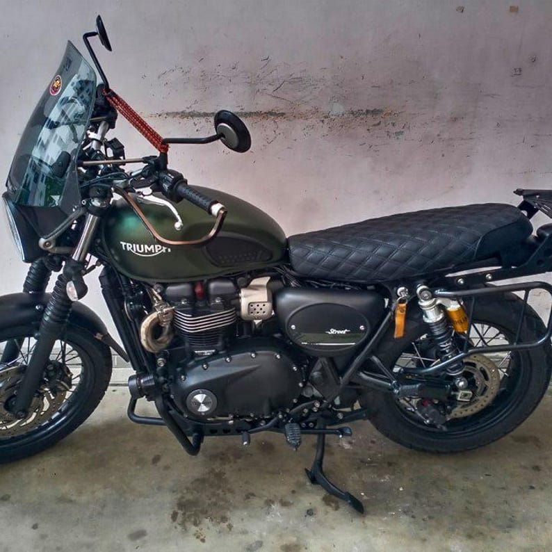 May include: A dark green Triumph Street motorcycle with a black quilted seat, black tires, and a clear windshield. The motorcycle has a black engine and frame, with gold-colored suspension components. The Triumph logo is visible on the fuel tank.