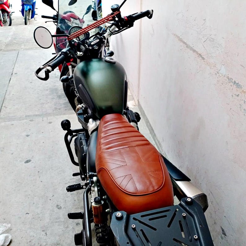 May include: A dark green motorcycle with a brown leather seat featuring a Union Jack design. The motorcycle has black handlebars, a round side mirror, and a black luggage rack. Other motorcycles are visible in the background.