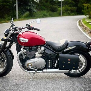 May include: A red Triumph motorcycle with a black leather saddlebag. The motorcycle has a chrome exhaust and a chrome engine. The motorcycle is parked on a paved road.