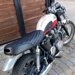 May include: A black motorcycle seat with a quilted pattern, mounted on a white and red motorcycle with a chrome exhaust pipe. The motorcycle has a black leather seat and a chrome exhaust pipe.