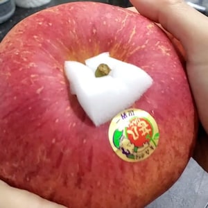 Könnte beinhalten: Ein roter Apfel mit einem weißen Aufkleber an der Seite. Der Aufkleber enthält japanischen Text und ein kleines grünes Blatt. Ein weißes Wattepad bedeckt den Stiel des Apfels.