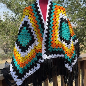 May include: A colorful, handmade crochet poncho with a geometric pattern. The poncho features squares of turquoise, yellow, orange, black, and white, with long black fringe along the bottom edge. The garment is displayed on a mannequin.