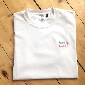 Puede incluir: Camiseta blanca con el texto "trans is beautiful" bordado en hilo rosa y azul.