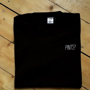 以下が含まれることがあります： 白い刺繍で「PINTS？」と書かれた黒いTシャツ。首元には「DOON DUH VAR」と書かれた小さなタグが付いています。木製の表面に折りたたまれたシャツ。