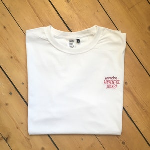 May include: A white t-shirt with the embroidered text "wannabe APPRENTICE JOCKEY" in red. The collar features a tag with "DOON DUR VAIL". The shirt is neatly folded on a wooden surface with diagonal planks.