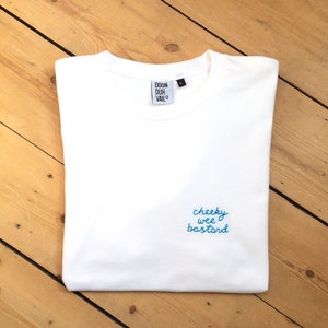 Può includere: Maglietta bianca con testo ricamato in blu che recita "cheeky wee bastard" e un'etichetta nera che recita "Doon Duh Vail Co."