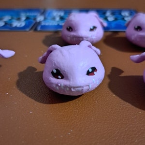 May include: Five small pink figurines with black eyes and red pupils. The figurines have a cartoonish appearance and are made of a soft material.