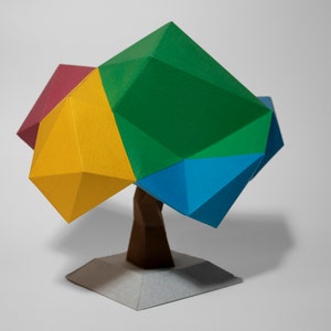 May include: A colorful papercraft tree sculpture with a green, blue, yellow, and red geometric canopy and a brown paper trunk. The tree is standing on a white paper base.