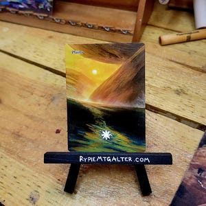 May include: A Magic: The Gathering card titled "Plains" is displayed on a miniature black easel. The card features a landscape with a bright sun setting over mountains. The easel has the text "RYPIEMTGALTER.COM" printed on it.