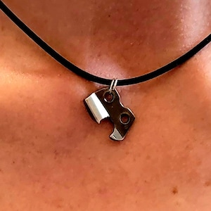 May include: A silver necklace with a pendant shaped like a puzzle piece. The necklace is made of black cord and is worn by a person.