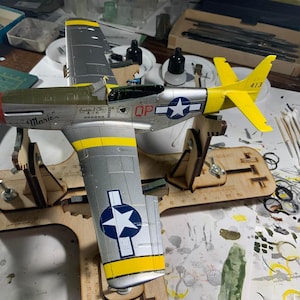 Könnte beinhalten: Ein Modellflugzeug, silberfarben mit gelben und olivgrünen Akzenten, ist auf einem Holzständer montiert. Das Flugzeug hat ein Sternabzeichen und den Namen "Marie" auf der Seite. Das Heck ist gelb mit der Nummer 413.