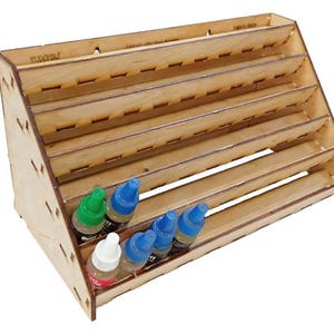May include: A wooden paint bottle organizer with multiple shelves. The organizer is angled for easy access and holds several bottles of paint in various colors. The wood has a natural finish, and the organizer is designed to hold small bottles.