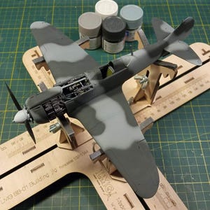Könnte beinhalten: Ein detailliertes Modellflugzeug in Grau- und Grüntönen mit Tarnmuster. Das Modell hat einen Propeller und ein offenes Triebwerk. Es steht auf einer Holzvorrichtung mit Zentimeter-Markierungen.