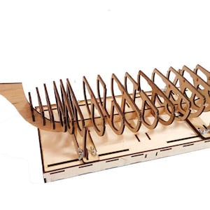 Op de afbeelding: Een houten modelbouwpakket voor een schip met een gedetailleerde romp en dek. De kit wordt gemonteerd op een houten basis en bevat een verscheidenheid aan kleine onderdelen.