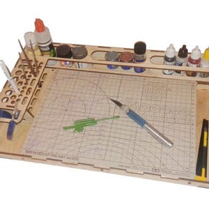 Może przedstawiać: Drewniane stanowisko do modelarstwa z matą do cięcia, butelkami z farbą i uchwytami na narzędzia. Stanowisko zawiera powierzchnię do cięcia z siatką, nóż modelarski i różne pojemniki na farby i materiały. Widoczny jest tekst "TAMIYA EXCUTTING MAT A4 SIZE".