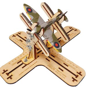 May include: A model Spitfire aircraft, painted in camouflage colours, is mounted on a wooden stand. The stand has a cross-shaped base with measurement markings. The plane has a propeller and red and yellow accents.