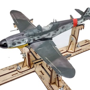 Może przedstawiać: Model samolotu, pomalowany w kamuflaż, jest zamontowany na drewnianym stojaku. Stojak ma linijkę i oznaczenia do precyzyjnego ustawienia. Samolot to Messerschmitt Bf 109, niemiecki myśliwiec z II wojny światowej.