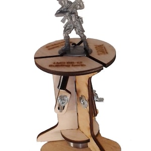 May include: A wooden rotating display stand for miniature figures. The stand has a circular base with three legs and a rotating platform. The platform has a small metal bearing in the center. The stand is labeled "LMG BB-12 Building Berth".