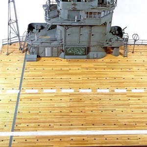 May include: A scale model of a gray naval ship island structure positioned on a light brown wooden deck. The deck features white painted lines and dark circular details. The gray superstructure includes a small rectangular sign with a map graphic.
