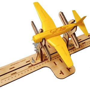 Könnte beinhalten: Ein gelbes Modellflugzeug, montiert auf einer Holzbasis mit Messmarkierungen. Das Flugzeug hat ein detailliertes Design, einschließlich Flügeln und einem Heck. Die Holzbasis hat Aussparungen und ist mit Metallbeschlägen befestigt.
