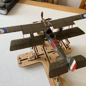 May include: A model of a vintage biplane with a brown and green paint scheme. The plane has a red and white stripe on the fuselage and a roundel on the wings. The plane is on a wooden stand with the text "IA" on the side.