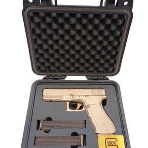 Puede incluir: Réplica de madera de una pistola, con dos cargadores y una placa dorada con la palabra "LOCK" dentro de una caja negra con acolchado de espuma. La caja es negra y tiene dos cierres.